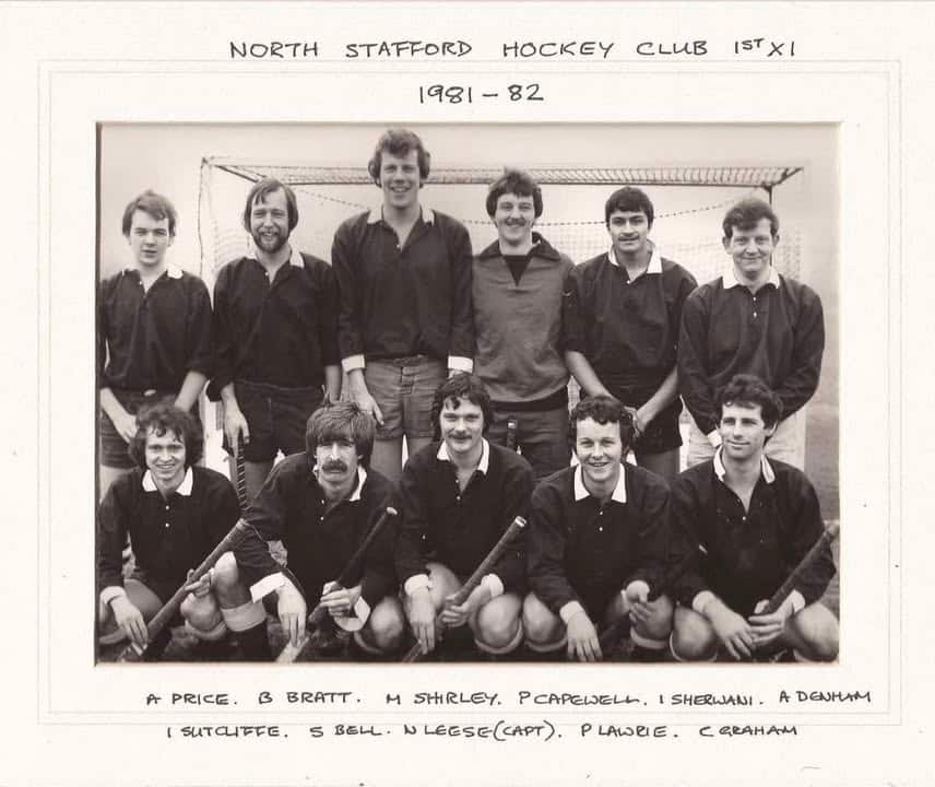 Black and white team photograph of young men in front of a hockey goal. They are wearing dark, long-sleeved hockey shirts. The back row standing and the front row is crouching.