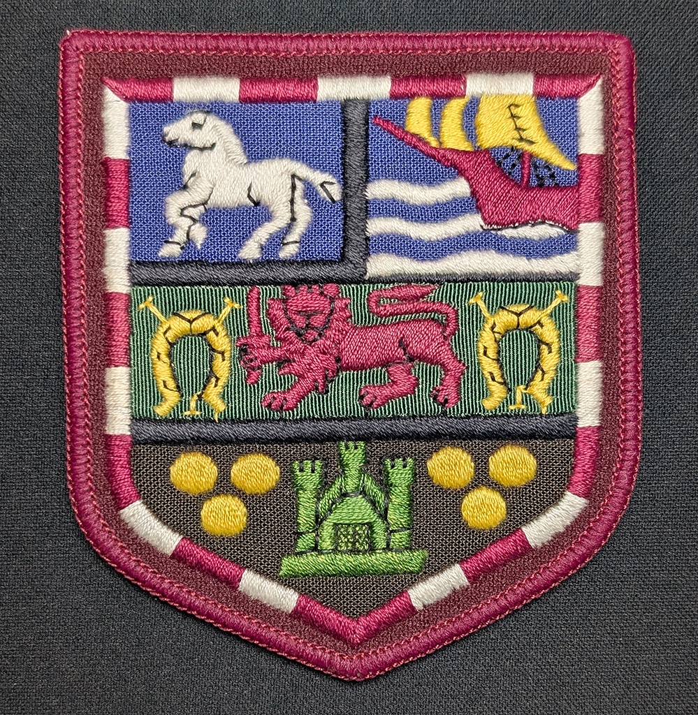 A multi-coloured cloth badge with red edging. The iconography includes a horse, a sail boat, a lion holding a sword and a castle.