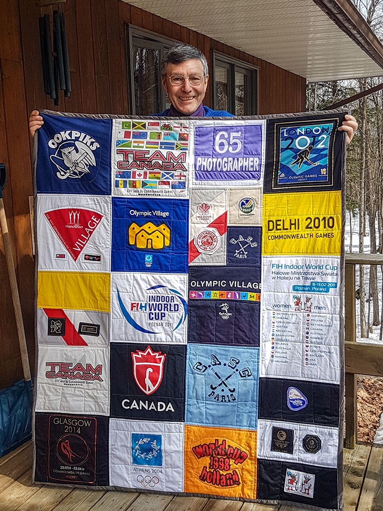 A man with grey hair stands on the veranda of a house holding up a large patchwork quilt.