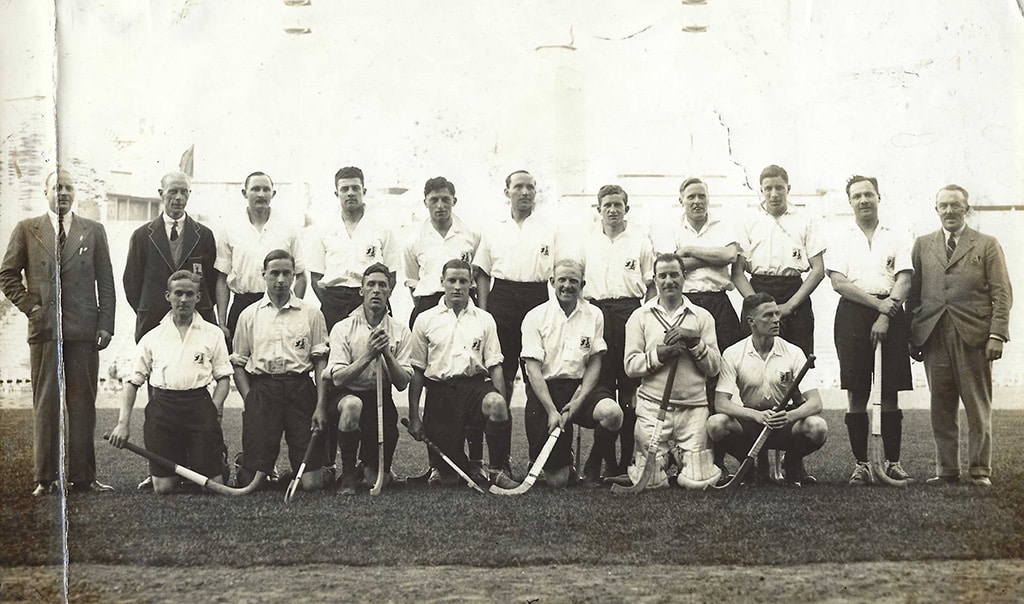 Black and white photograph of a men's hockey team in two rows, the back row standing and the front row kneeling. The men are wearing white shirts with badges of the islands of Great Britain and Ireland and long, knee-length shorts.