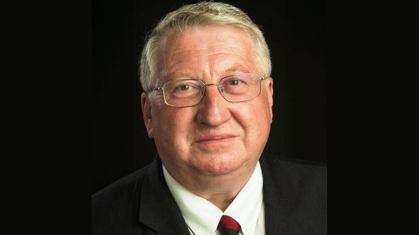 Photograph of a man in profile with short, blonde, grey-ing hair and glasses. He is wearing a blazer, shirt and red tie.