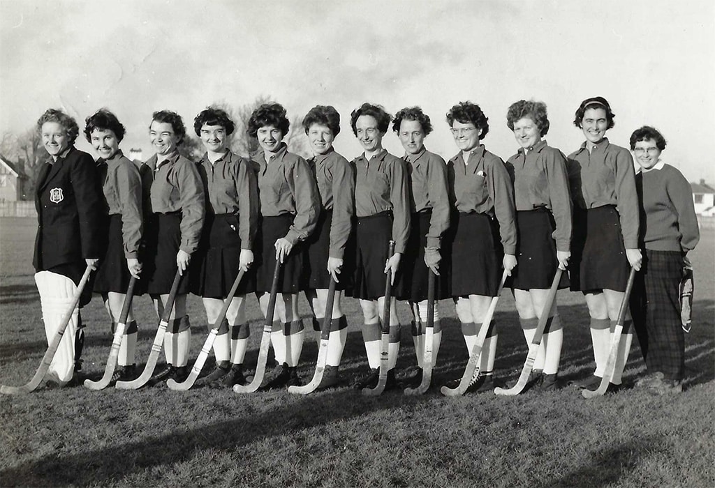 A row of standing women in skirts, long white socks and playing shirts holding hockey sticks.