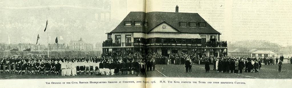 Black and white photograph of a two-storey pavilion in the middle of a sports ground. Large groups of people are gathered in front of the building.
