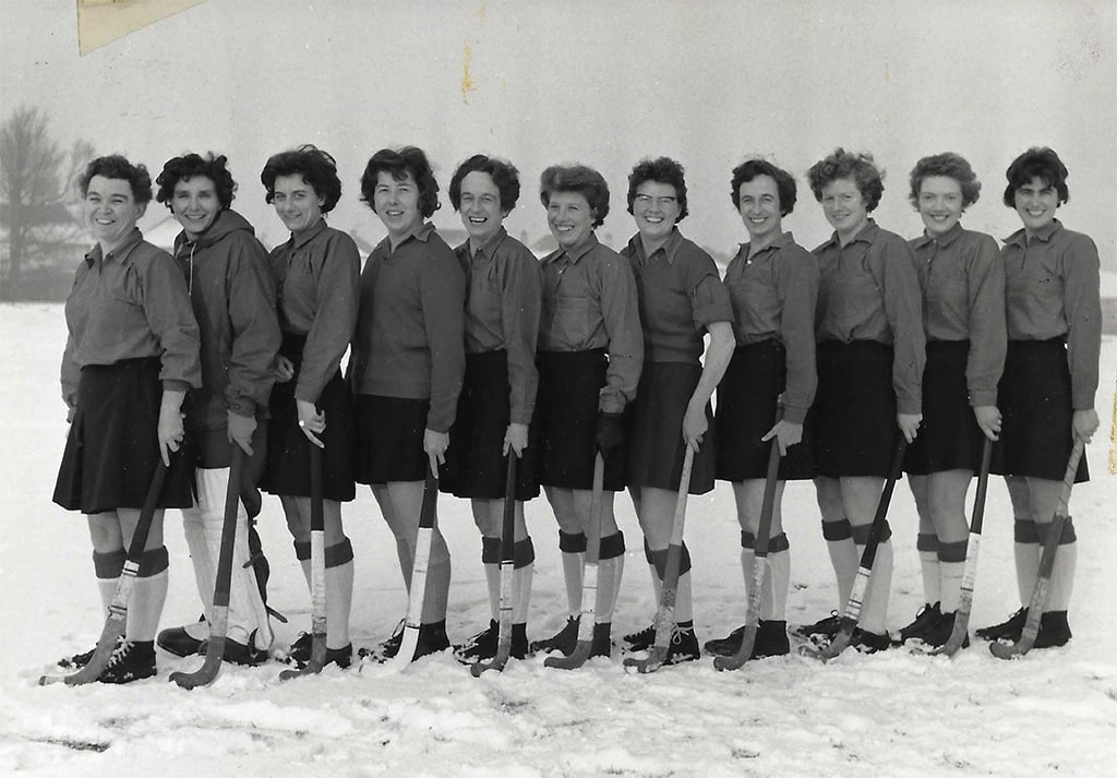 A row of standing women in skirts, long socks and playing shirts holding hockey sticks. The ground is covered in snow