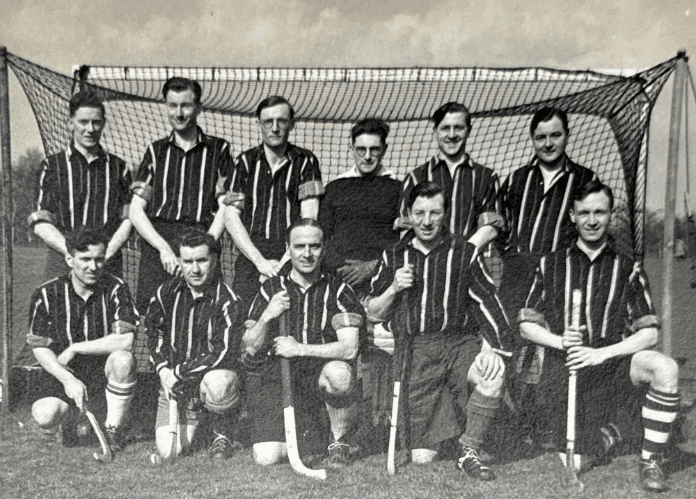 A team photo of men standing or kneeling in two rows in front of a goal. They are wearing striped playing shirts and holding hockey sticks.