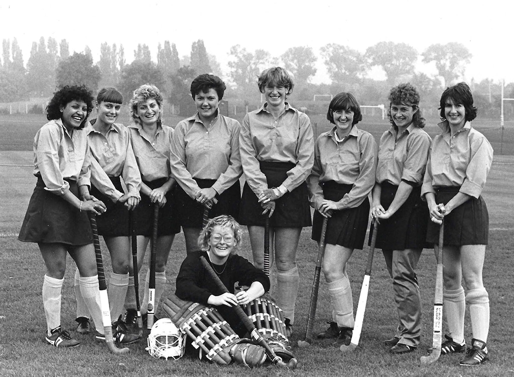 A row of standing women in skirts, long white socks and playing shirts holding hockey sticks. A goalkeeper in protective kit sits on the floor.