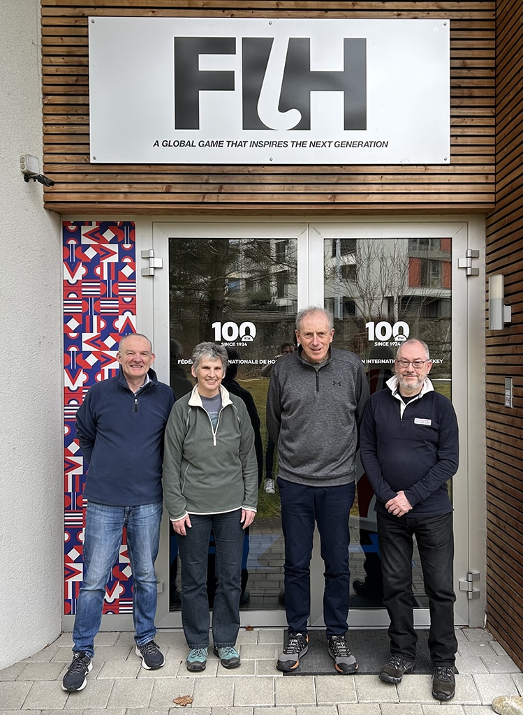 Three men and a woman stand before large doors. A large sign above the door bears the letters FIH
