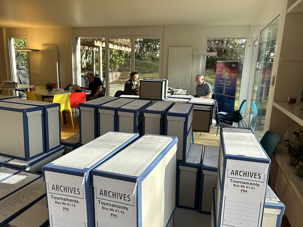 A well-lit room with large windows, archival boxes in the foreground and three people working at desks in the background