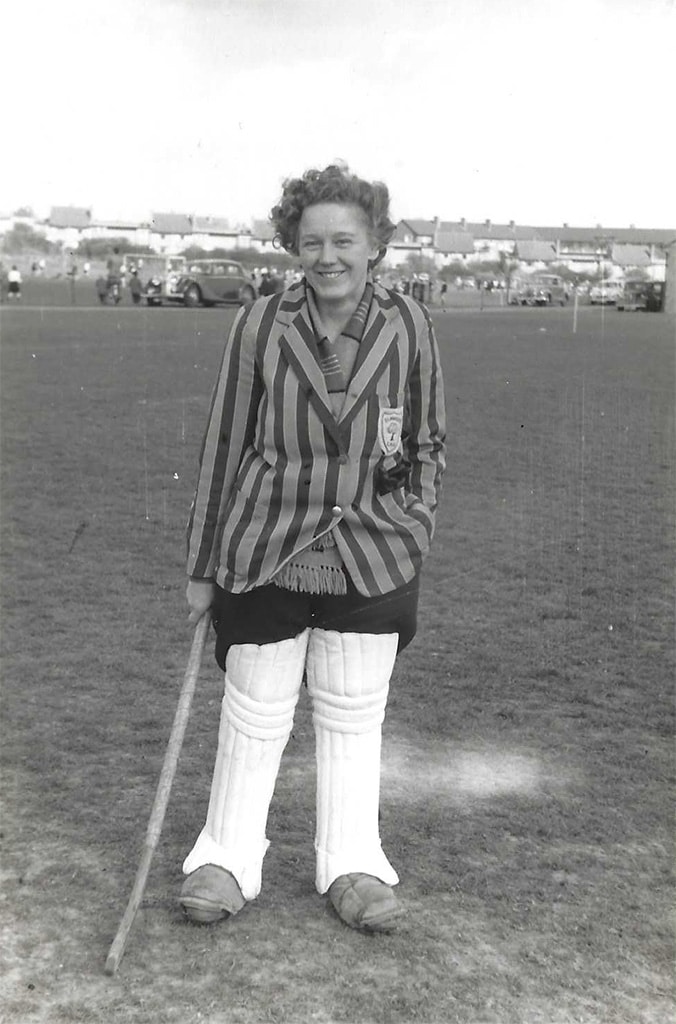 A woman wearing a striped blazer and cricket pads holds a hockey stick.