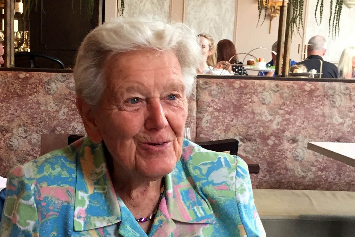 An elderly lady with white hair is seated wearing a blue and pink shirt.