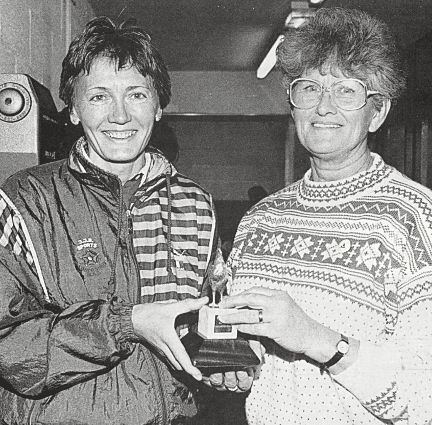 Two women holding a small trophy of a bird on a plinth. The woman on the left is younger and wearing a tracksuit. The women on the right is older and wearing a sweater and large spectacles.