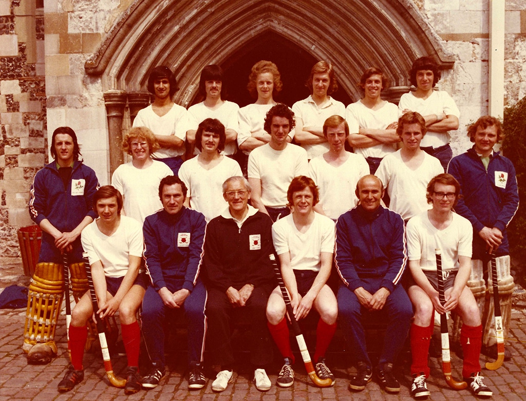 Three rows of young men wearing white shirts, dark shorts and red socks. Most have long hair. The front row is seated and the back two rows standing.