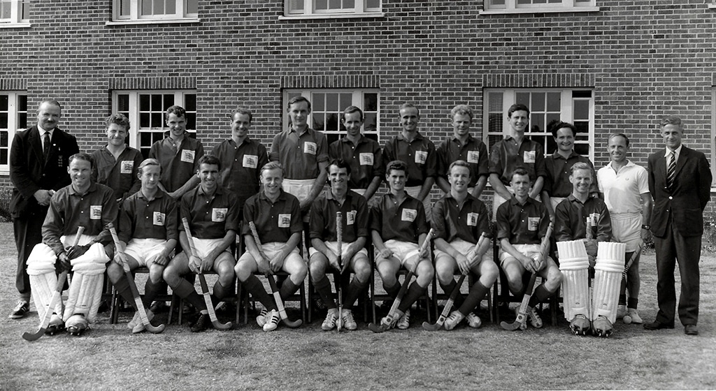 Black and white photograph of two rows of men holding hockey sticks, the front row seated and the back row standing. They are wearing dark shirts with Union flag badges and white shorts.