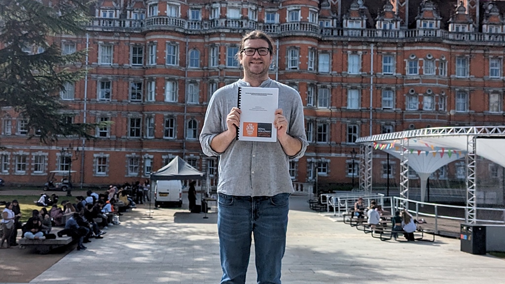 A young man in jeans and a grey shirt standing before a large, ornate red-brick building.
