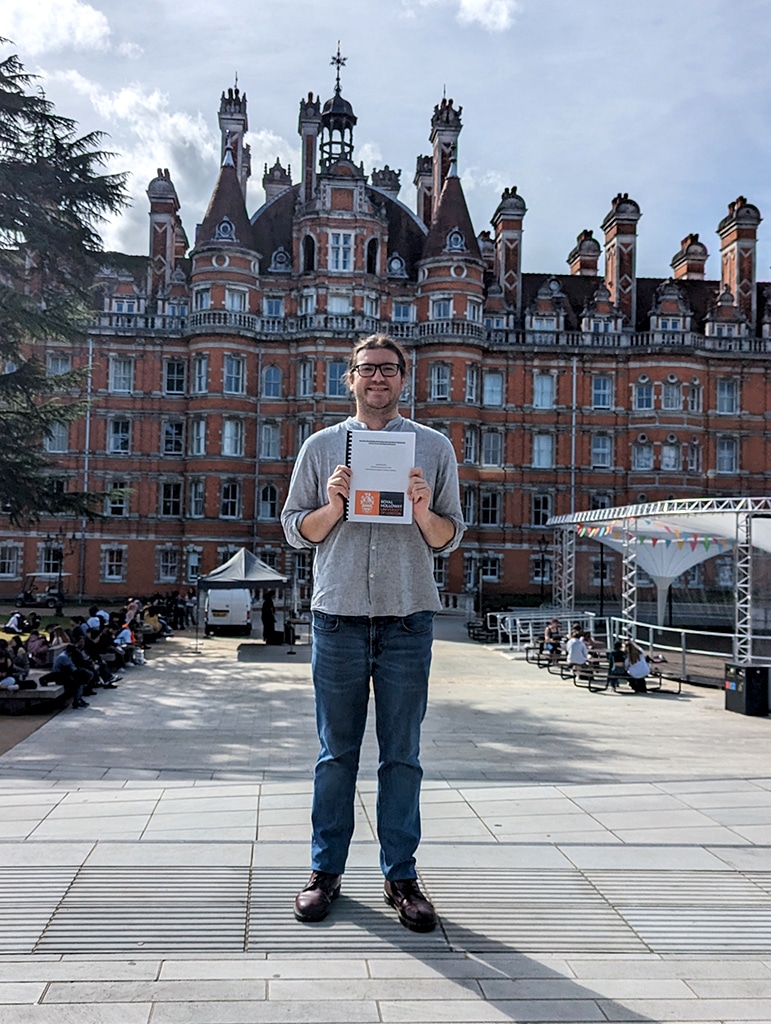 A young man in jeans and a grey shirt standing before a large, ornate red-brick building.