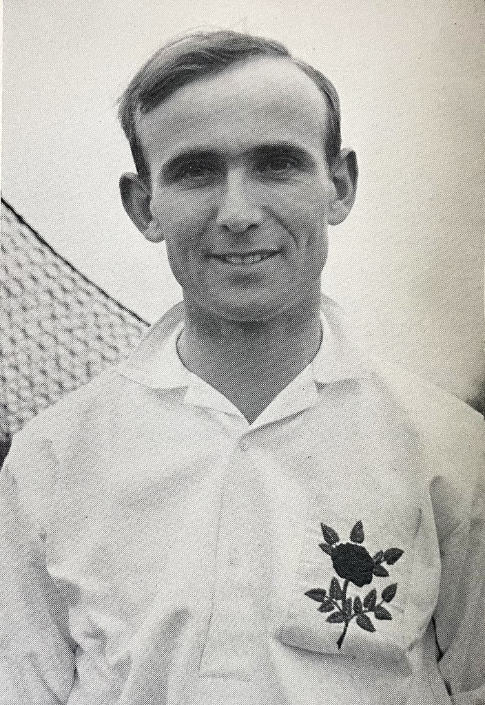 A middle-aged man wearing a white shirt with a rose on the chest.