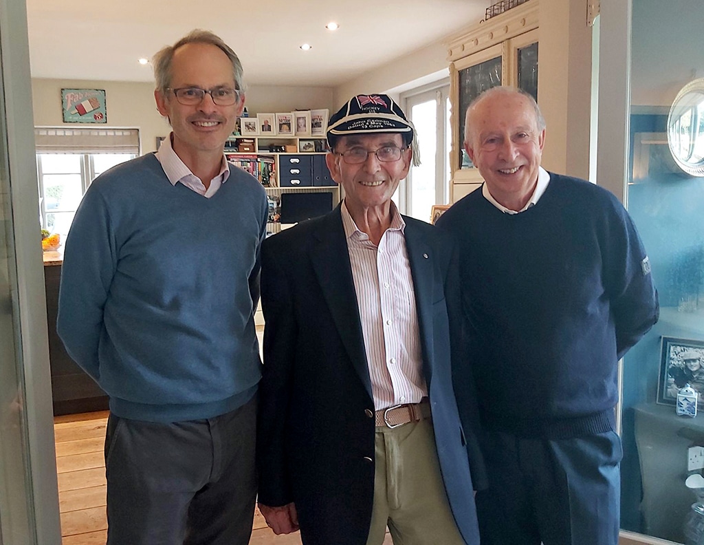 Three men smile at the camera: the middle man is wearing a navy blue cap and blazer