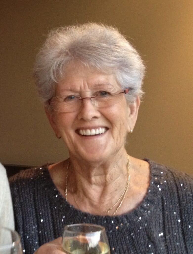 A smiling elderly lady with grey hair and glasses holds a glass of wine.
