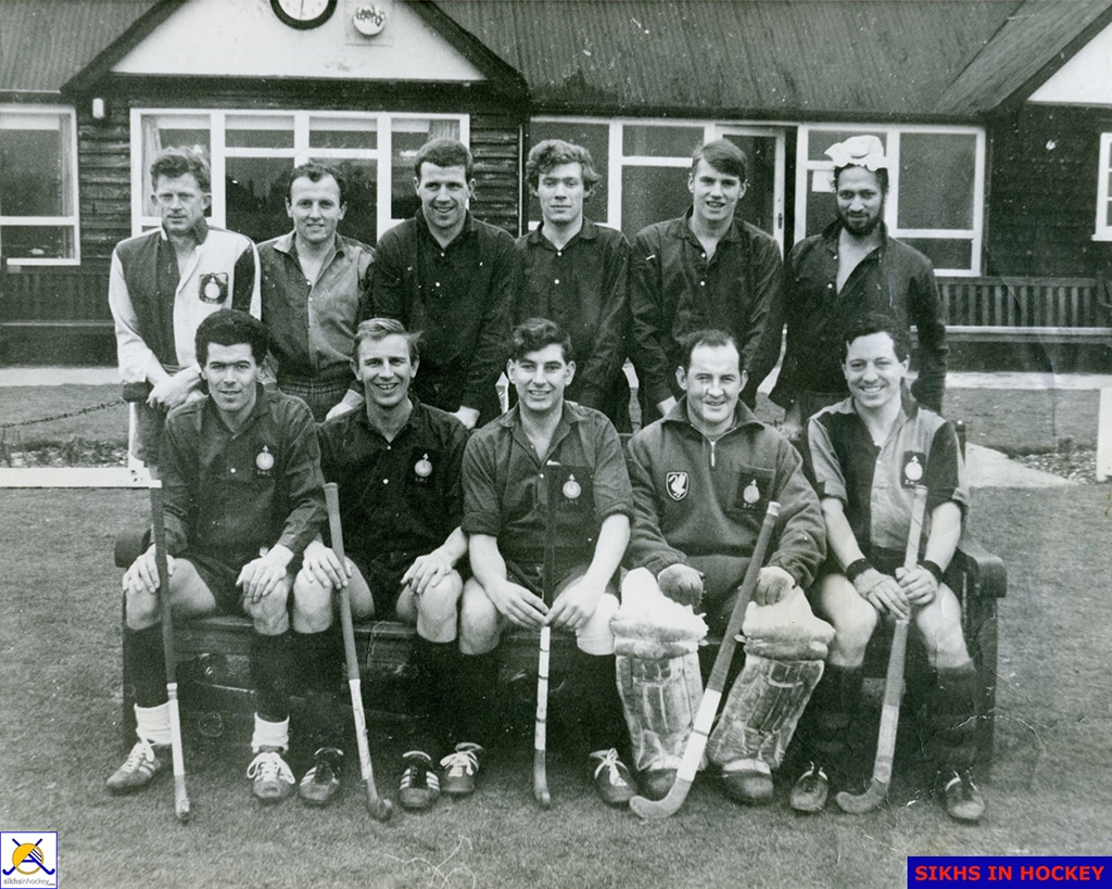 A hockey team of men standing and seated in two rows in front of a wooden clubhouse building. One player is South Asian, sporting a joora (topknot); the rest are Caucasian.