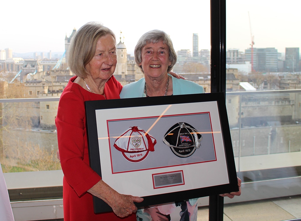 Two women holding a large frame within which are set two cloth caps. One cap is white with red trim and tassel, the other is navy blue with silver trim and tassel.