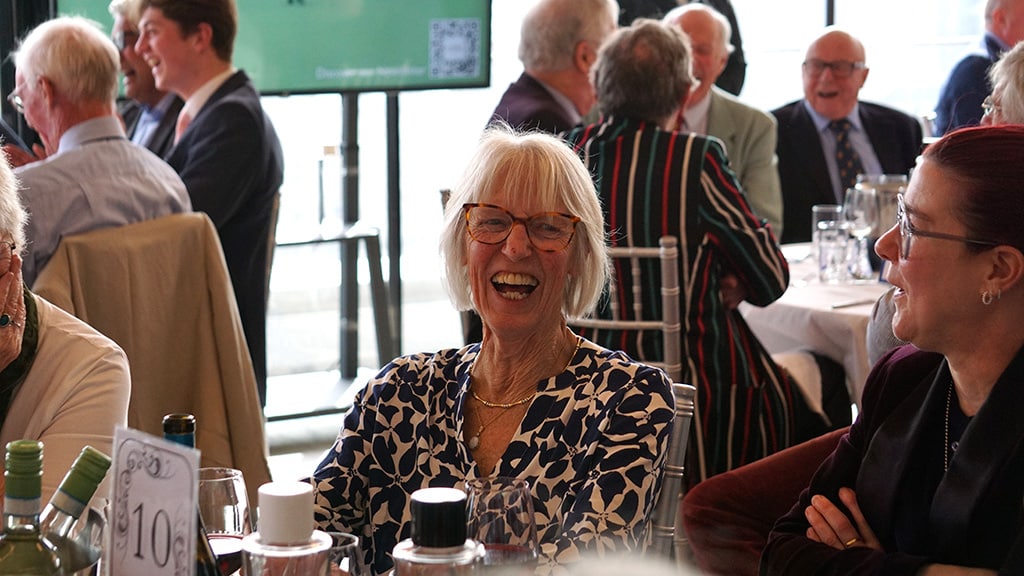 A woman laughs loudly in a room of similarly happy people seated at tables.