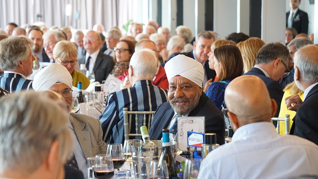A room of people seated at large round tables. A man wearing a turban smiles directly at the camera.