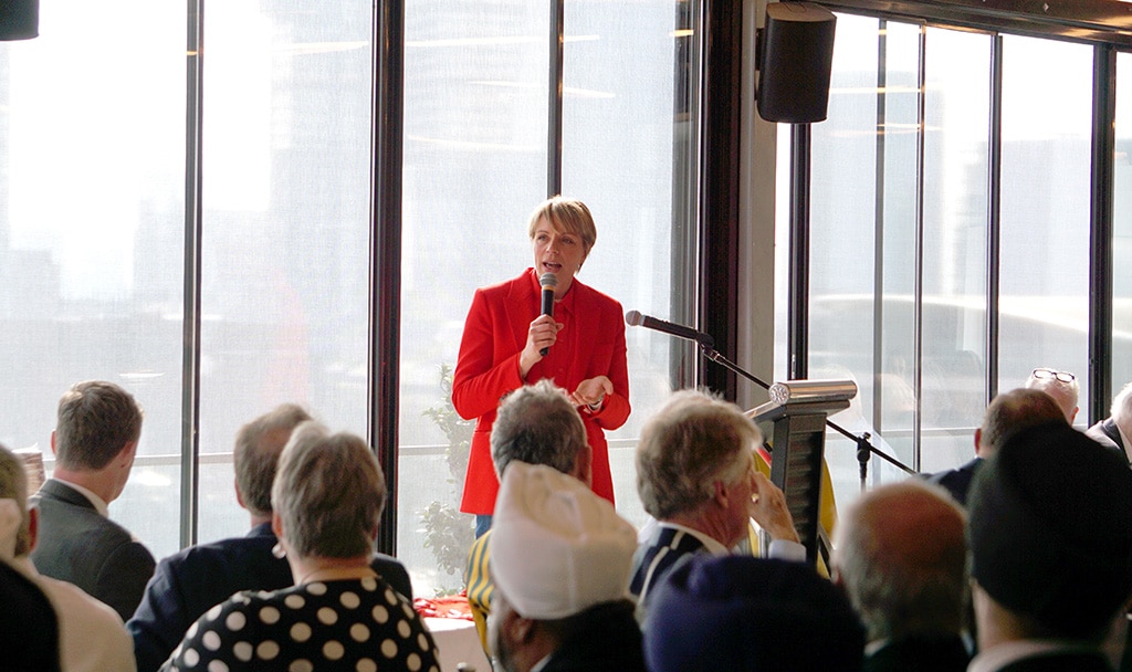 A woman in a red shirt and red blazer gives a speech to a large audience.