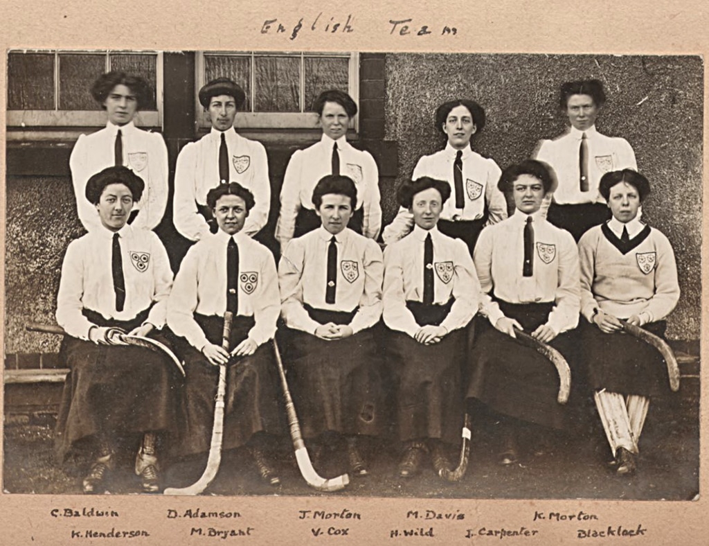 A photograph of two rows of women, the front row seated, wearing long skirts, white blouses and long ties. A cloth badge of three roses is on their breast pockets.