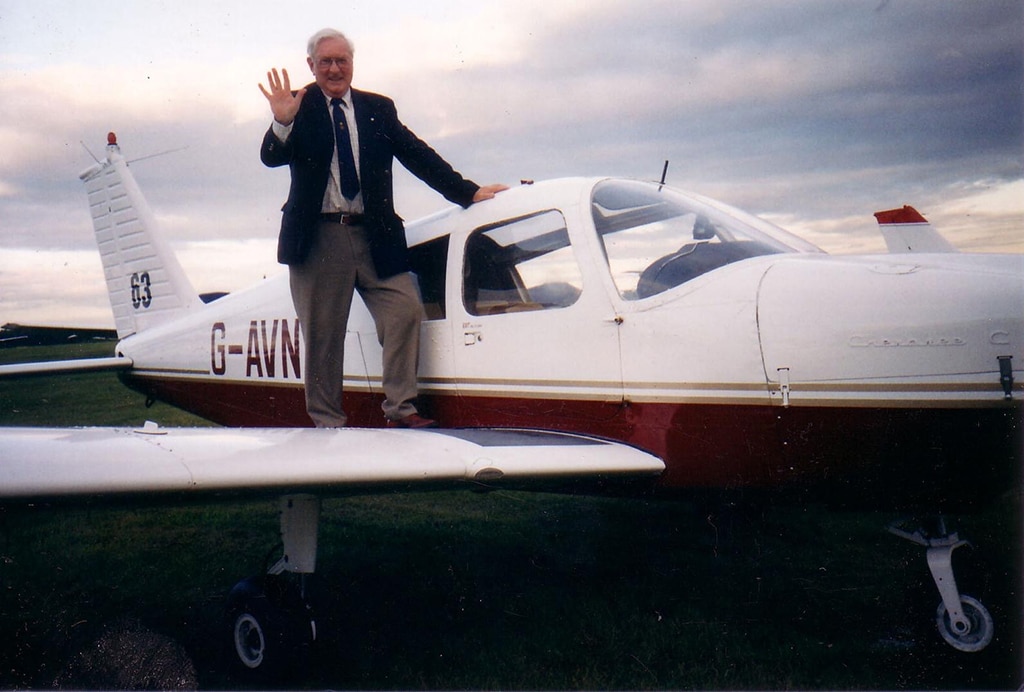 An elderly man wearing a blazer and tie stands on the wing of a small aeroplane and waves.