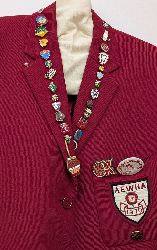 Detail of a red blazer adorned with many colourful pins on the lapel. The shield-shaped cloth badge on the blazer pocket features a red rose on a white background.