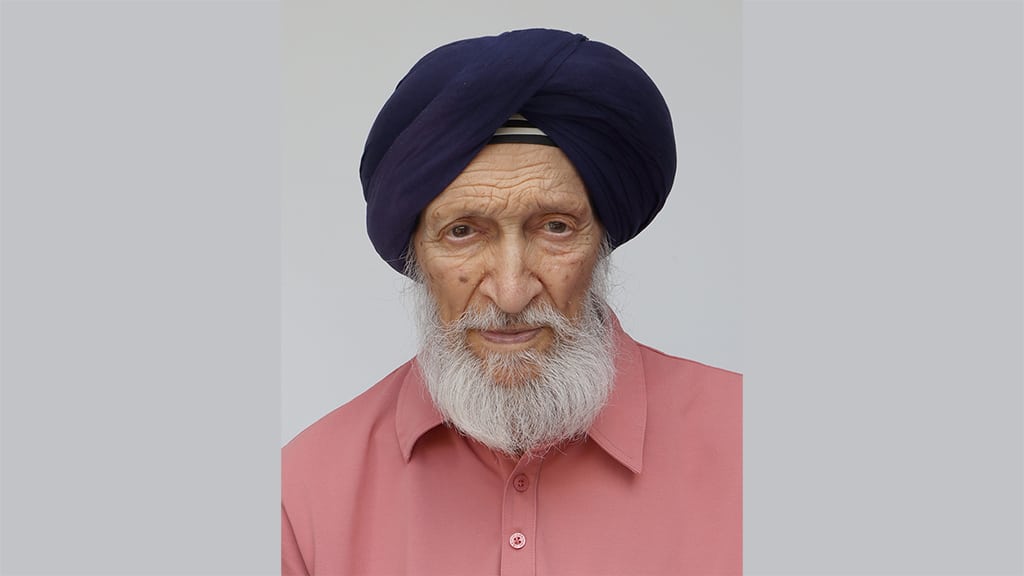 Portrait photograph of an elderly man of Indian heritage with a full grey beard, wearing a light pink shirt and navy blue turban.