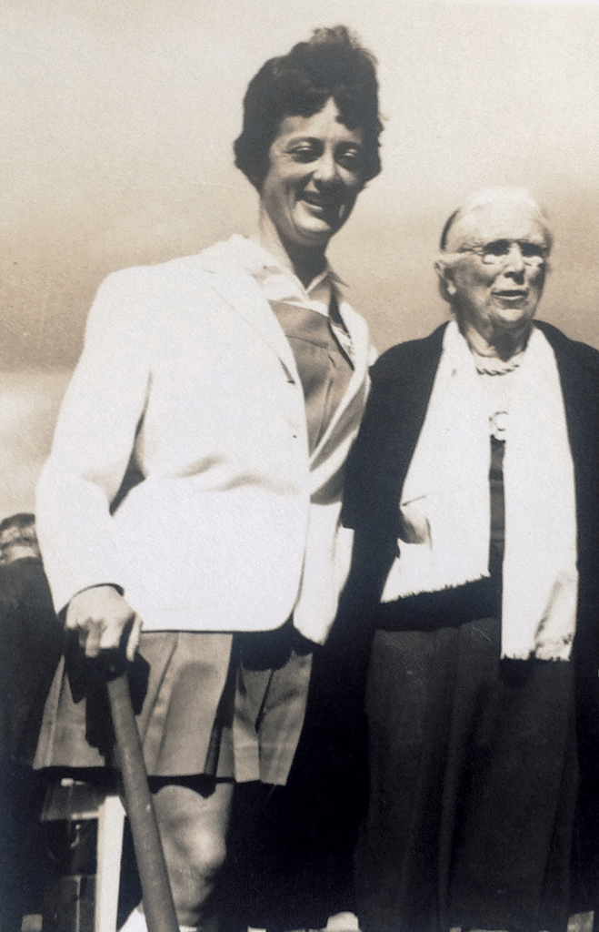 Black and white image of two women, one very elderly. The younger woman holds a hockey stick.