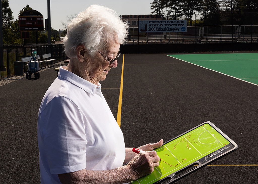 An elderly lady with grey hair and spectacles writes on a tactics board. A hockey pitch is in the background.