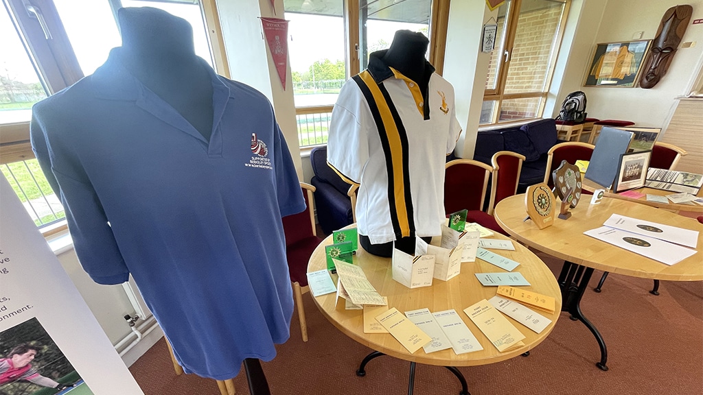 Two mannequins, one with a blue polo shirt and one with a white and yellow polo shirt and three round tables booklets, leaflets, shield trophies and photograph albums.