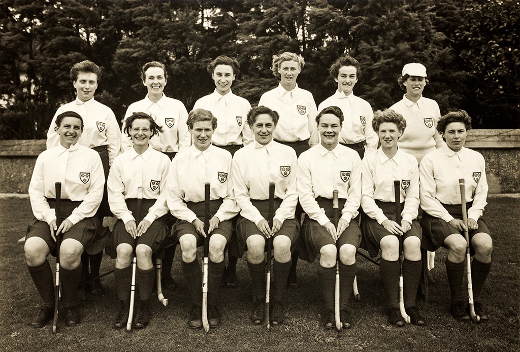 Black and white photograph of a smiling women's hockey team posed in two rows in front of trees, the back row standing and the front row seated holding hockey sticks. The players wear a shield-shaped badge of three roses on the breast pocket of their white button-up blouses.