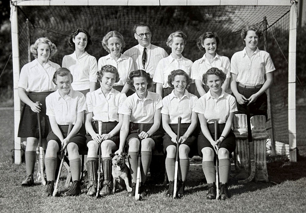Black and white photograph of a smiling women's hockey team posed in two rows in front of a goal, the back row standing and the front row seated holding hockey sticks.
