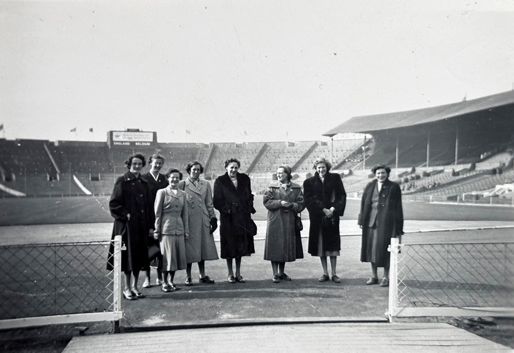 Black and white photograph of eight women in long coats stood in a line with a large empty stadium in the background.