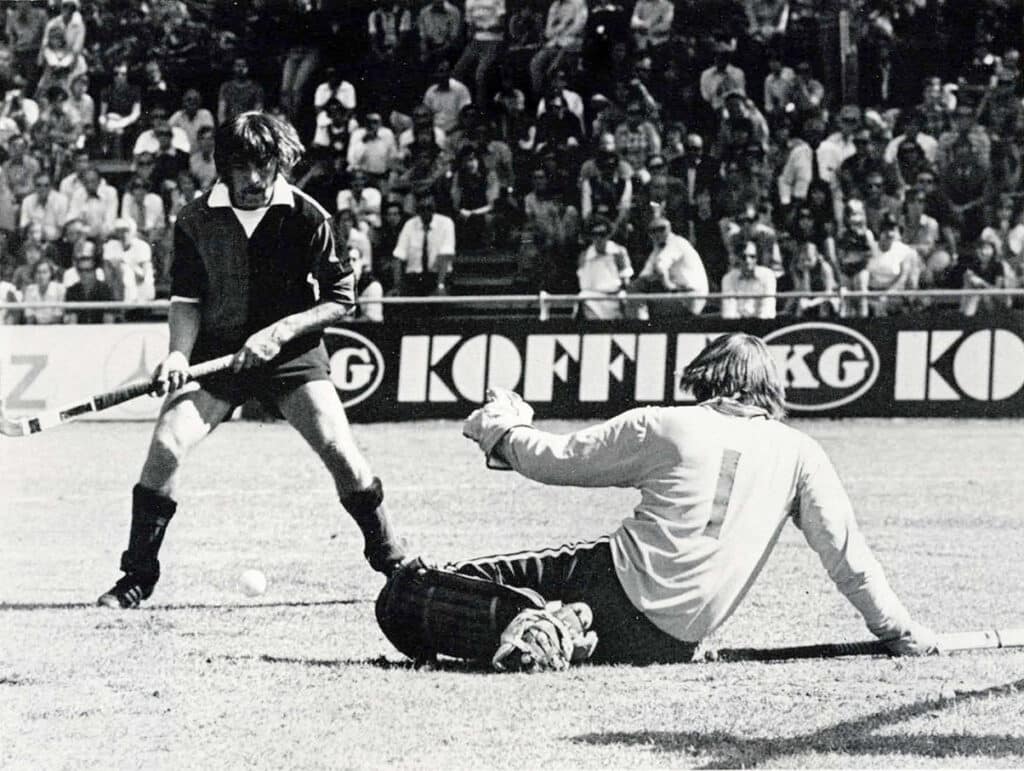 Black and white action photograph of a hockey match on grass. A goalkeeper spreads himself on the floor to save with a defender close behind. A crowd watches on.