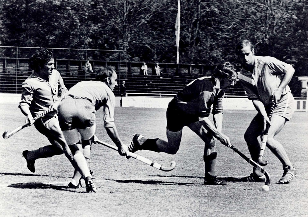 Black and white action photograph of a hockey match on grass. One man dribbles the ball between three defenders.