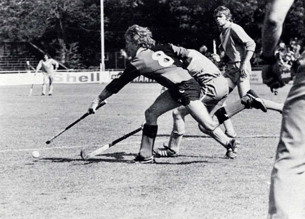 Black and white action photograph of a hockey match on grass. Two players compete aggressively for a loose ball.