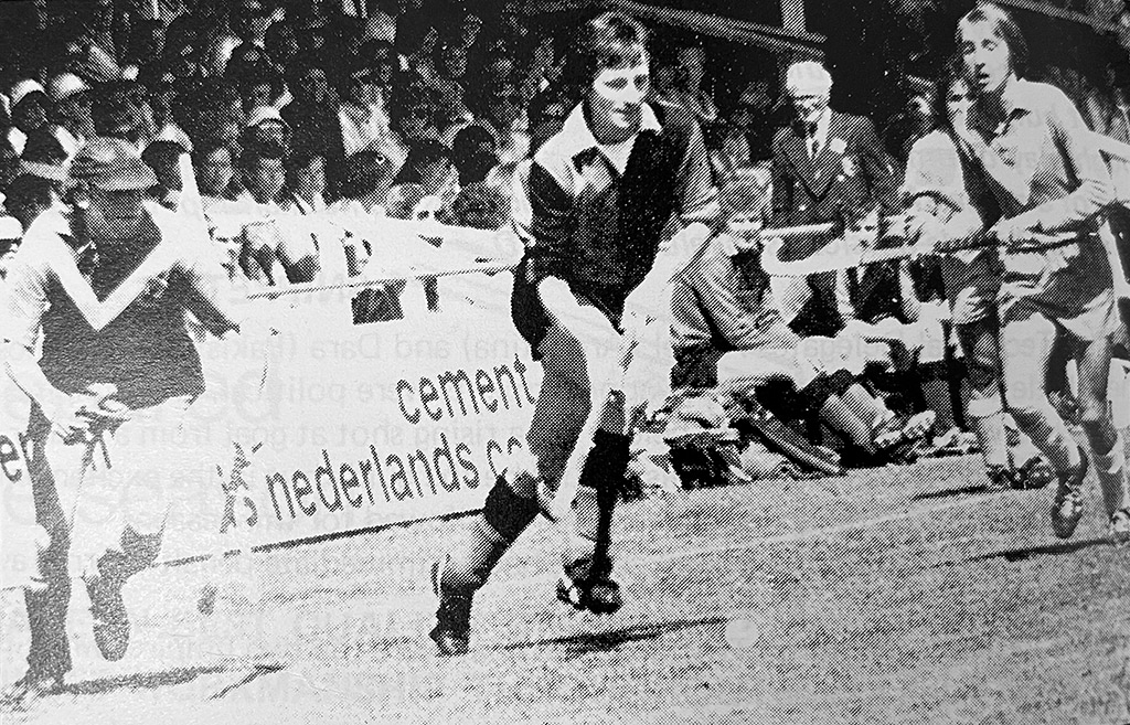 Black and white action photograph of a hockey match on grass. Players run towards camera with a crowd behind.