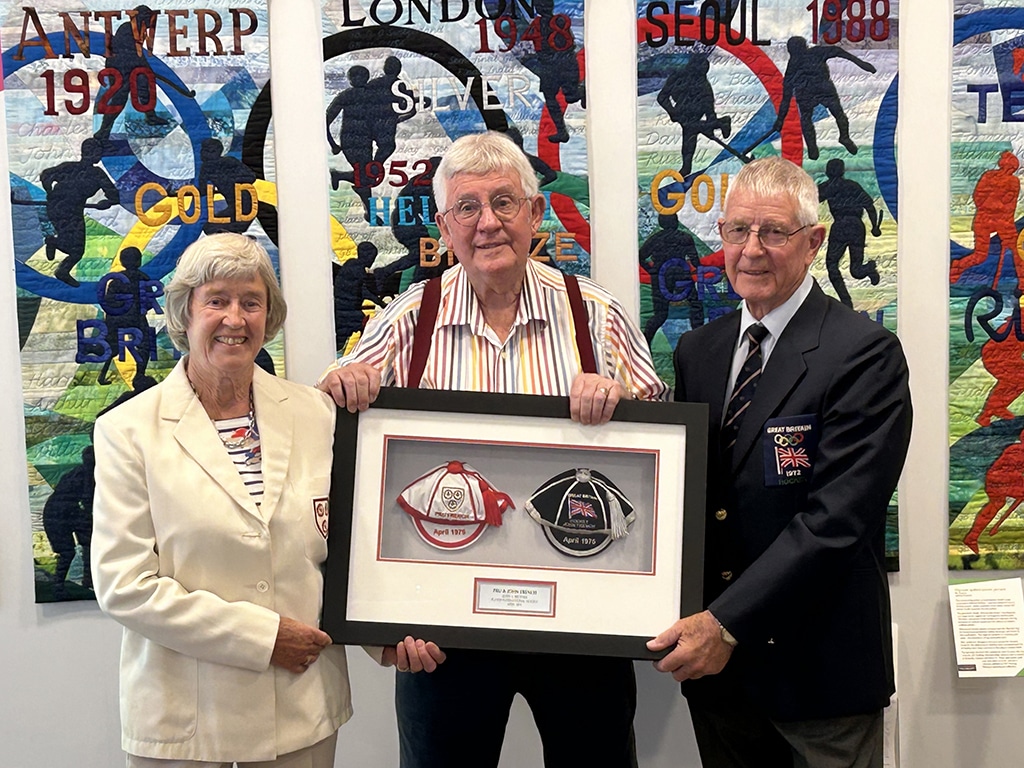 Three elderly people pose for a photograph in a museum exhibition hall holding a large frame with two caps inside.