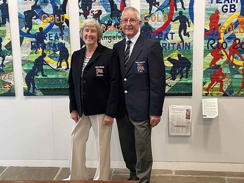 A man and a woman pose, smiling, wearing matching navy blue blazers standing before a colourful tapestry.