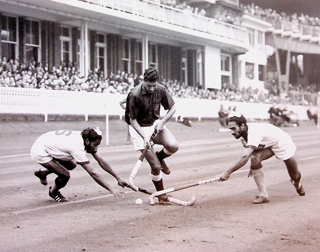 Black and white action photograph of a hockey match. Two men attempt to tackle another player dribbling with the ball towards camera. A large pavilion is in the background.