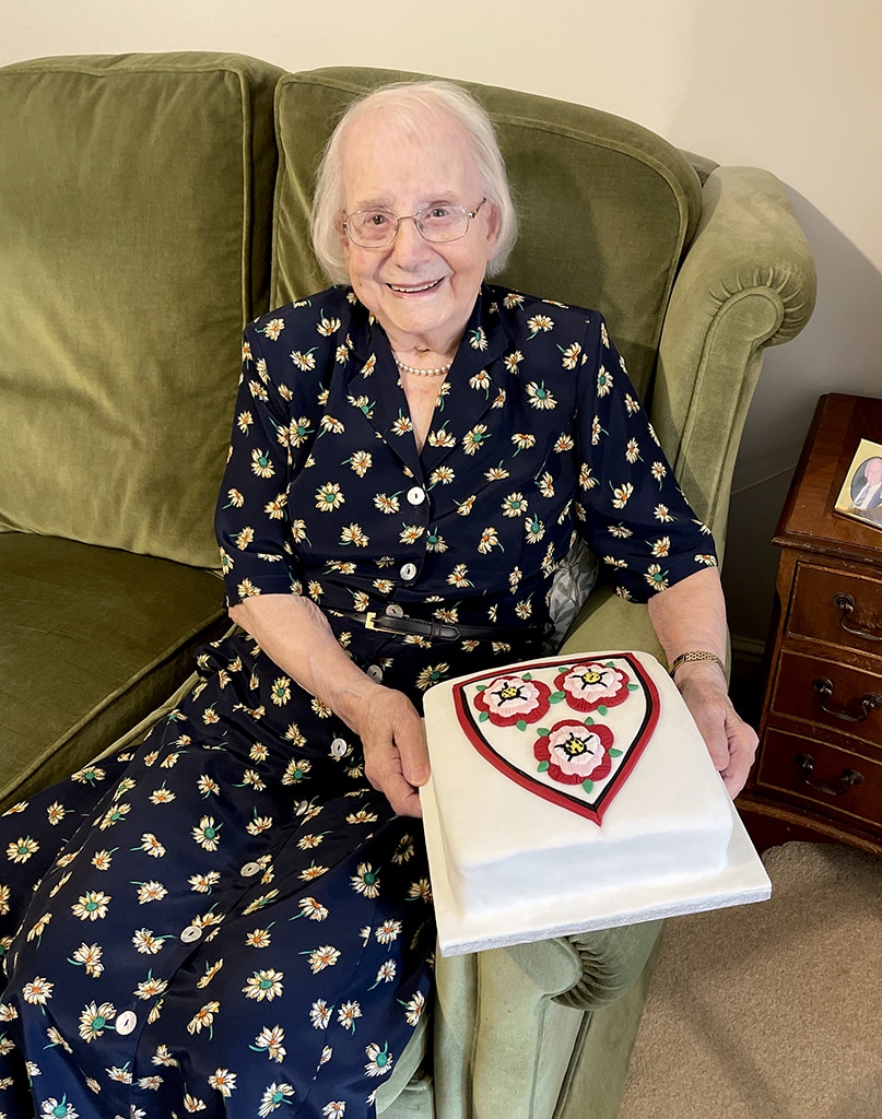 An elderly lady wearing a flowery dress sitting on a green sofa holding a large, square cake decorated with three roses within a shield.