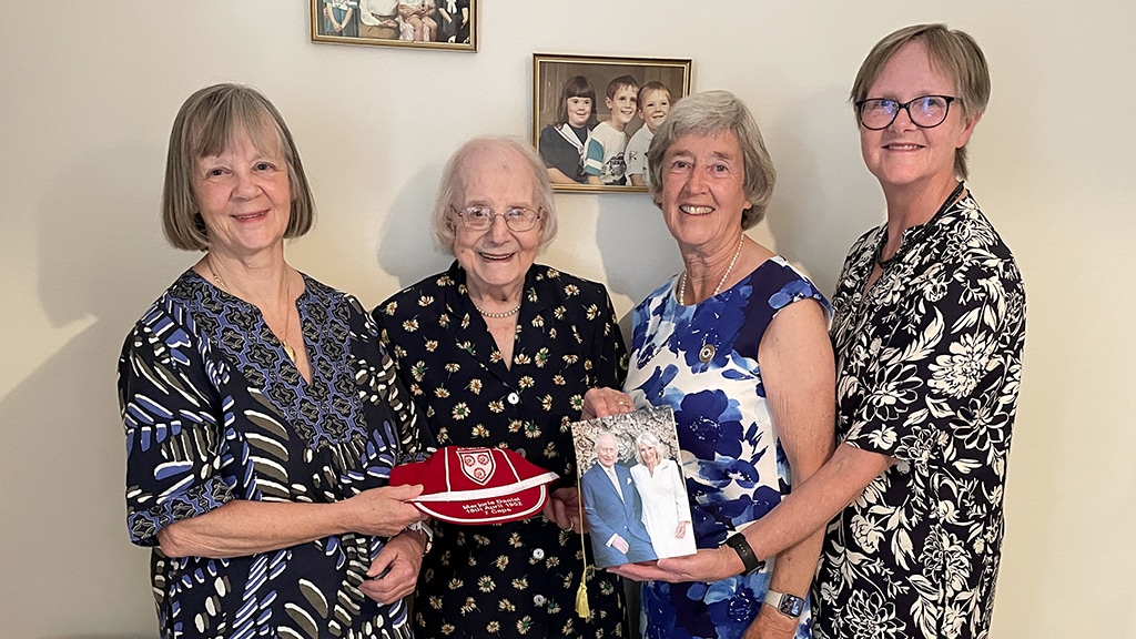 An elderly woman stands with three other women in a line holding a red and white embroidered cap and a large birthday card between them.