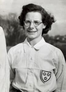 Profile photograph of a women with wavy brown hair and thick-rimmed spectacles wearing a button-up white blouse with a shield-shaped badge on three roses her breast.