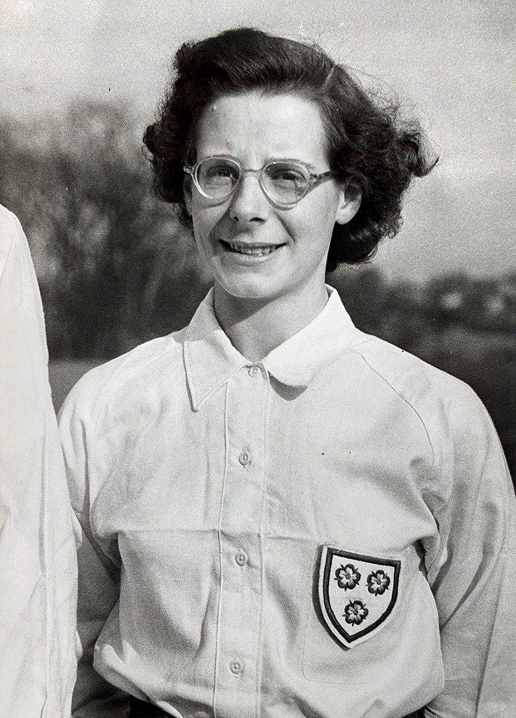 Profile photograph of a women with wavy brown hair and thick-rimmed spectacles wearing a button-up white blouse with a shield-shaped badge on three roses her breast.