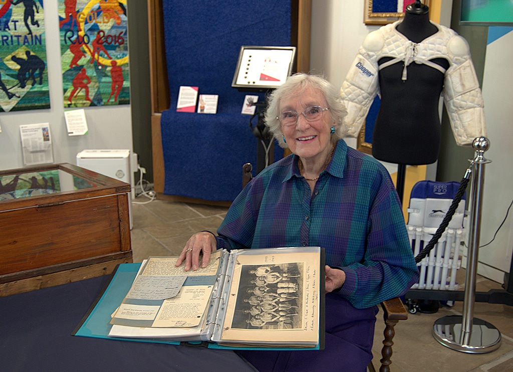 A smiling elderly lady dressed in blue and purple sits at a table holding open a scrapbook showing photographs and paper documents.