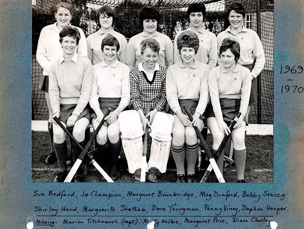 Black and white photograph of a smiling women's hockey team posed in two rows in front of a goal, the back row standing and the front row are seated holding hockey sticks crossed below their knees. The photograph is mounted on blue card.